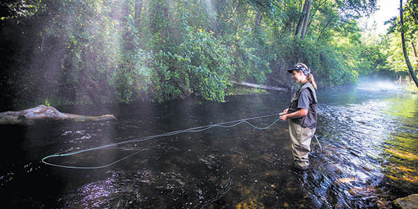 A woman wearing waders smiles as she fly fishes in a stream.
