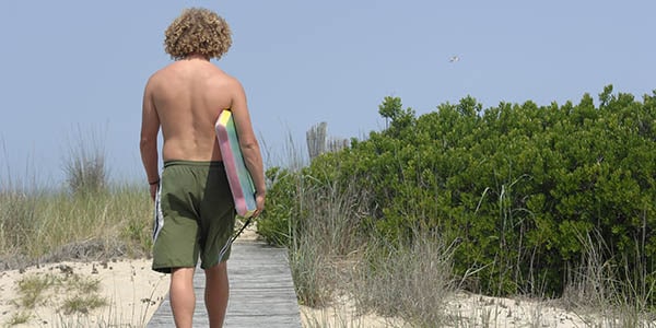 Curly-haired guy in bathing suit totes a boogie board along a wooden walkway toward the beach.