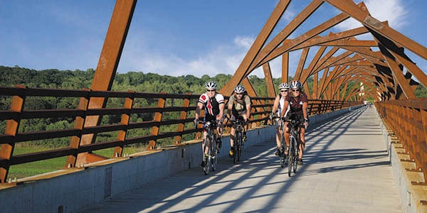 Four cyclists with helmets ride on a bridge with an interesting canopy.