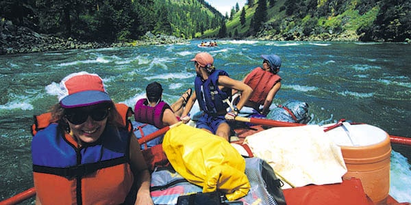 Four rafters wearing life vests approach a series of rapids in a river valley.