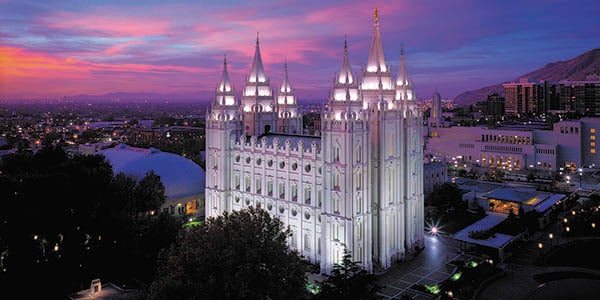 The Mormon Temple with six spires illuminated in the midst of a city.