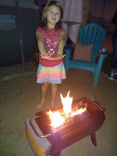 Young girl standing alongside the BioLite Firepit, warming her hands over the fire at the beach.