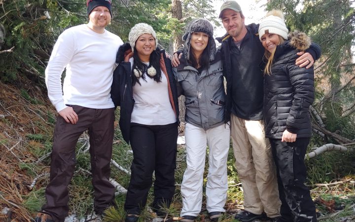 Five adults in snow clothing with snow and pine trees behind them.
