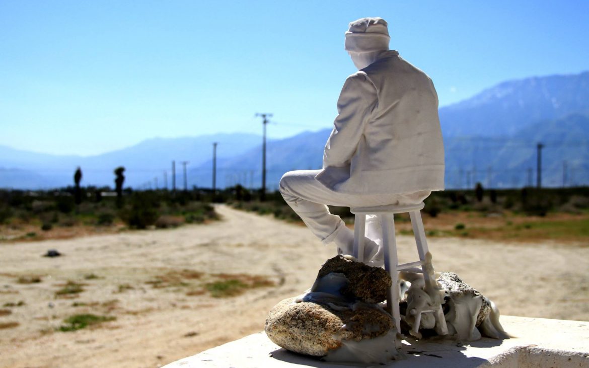 White sculpture of man on stool in the California desert.