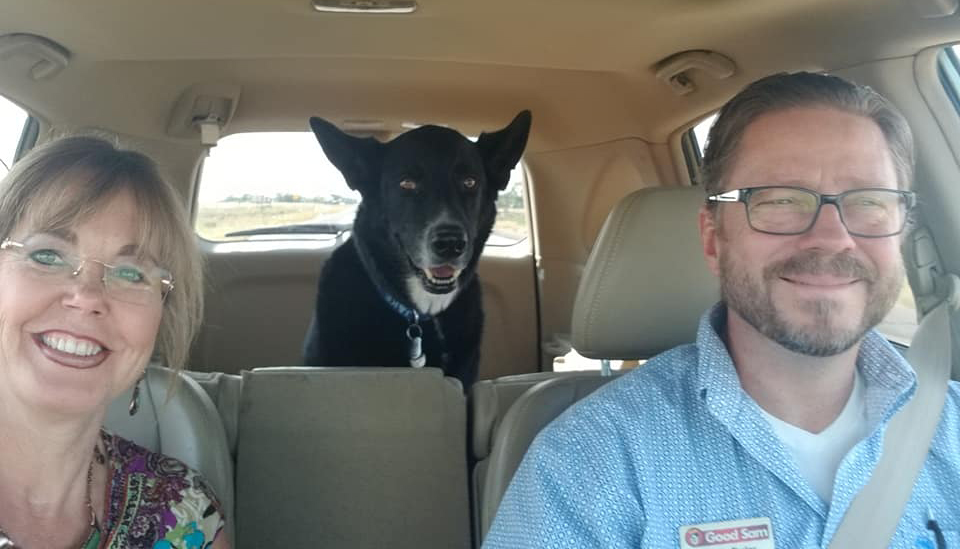 A smiling couple in vehicle with a dog in the back seat.