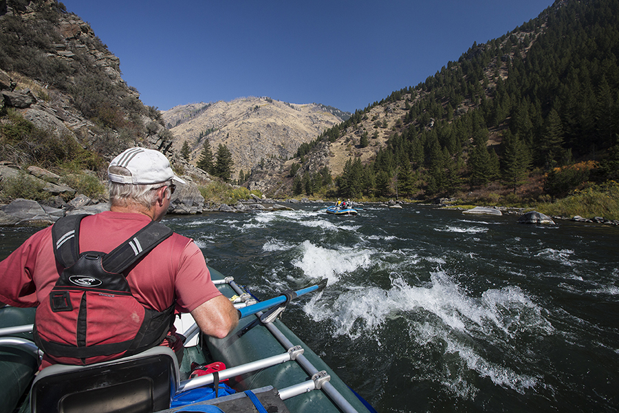 Using oars, a man guides his boat through whitewater in Montana with mountains in the background.