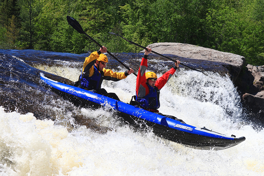 Riders of a blue kayak raise their oars in jubilation as their vessel plummets down gushing waterfalls on the Penobscot River. 
