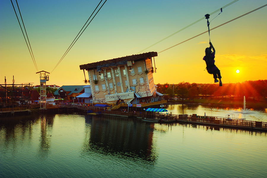 A zipline zooms across a lake with the Wonder Works building in the background.