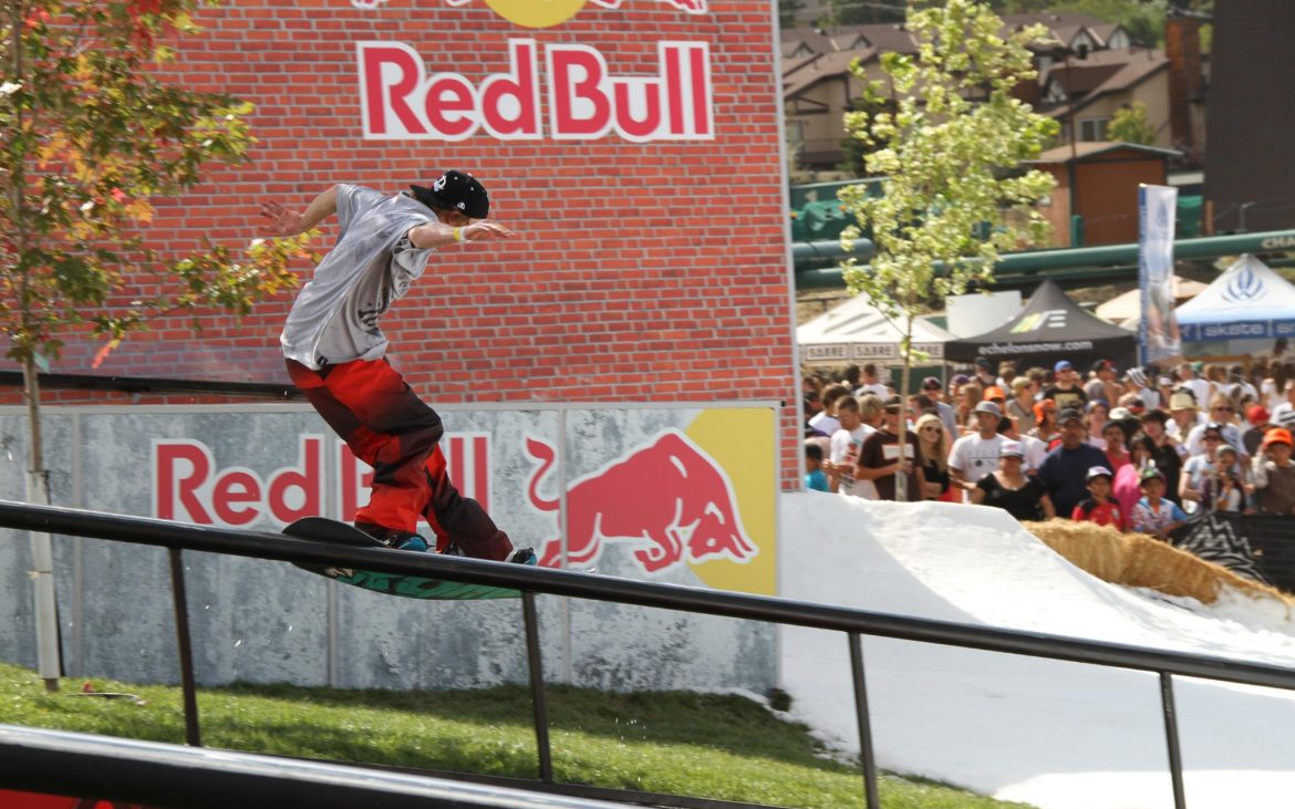 Young man snowboarding on rail with brick building and Red Bull logo in background
