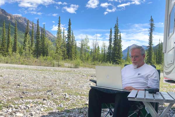Jim Nelson works on his laptop in British Columbia with rugged trees and mountains in the distance.