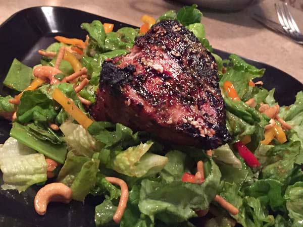 A grilled salmon steak sits atop a bed of greens, cashews and noodles
