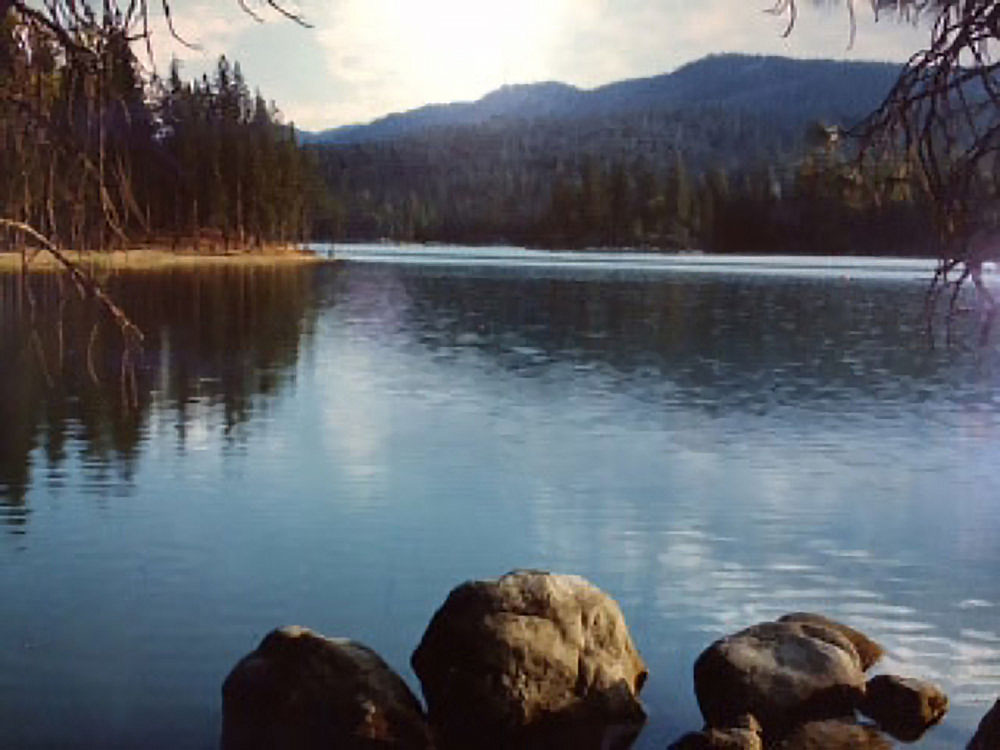 A lake reflects the sunny sky as forested mountains rise in the distance.