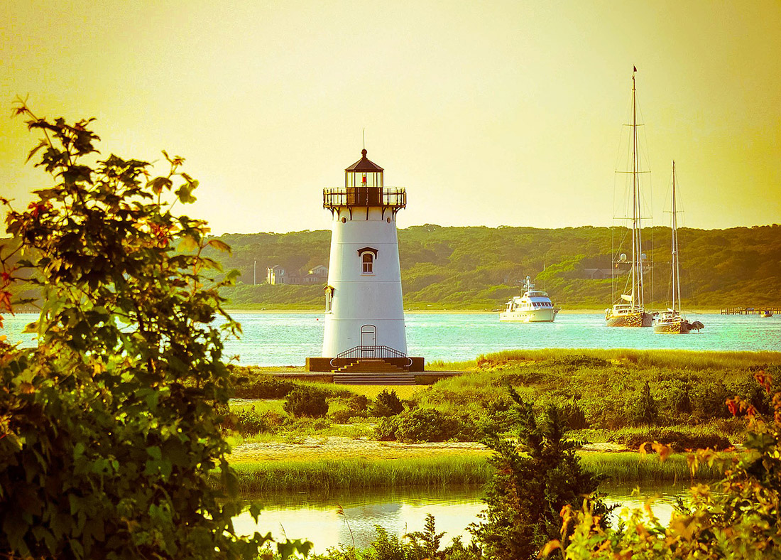A white lighthouse towers over an inlet with two sailboats and one cruising boat on the water in the background.