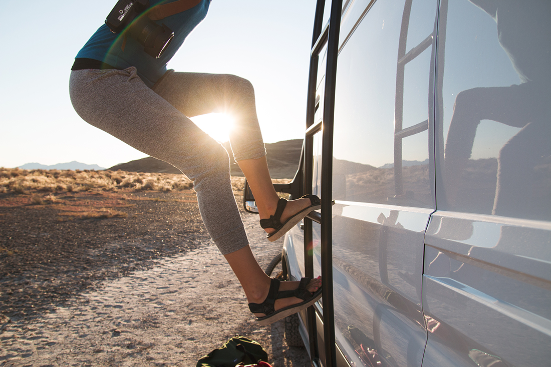 A woman climbs a ladder on the side of a white camper van as the sun rises in the distance.