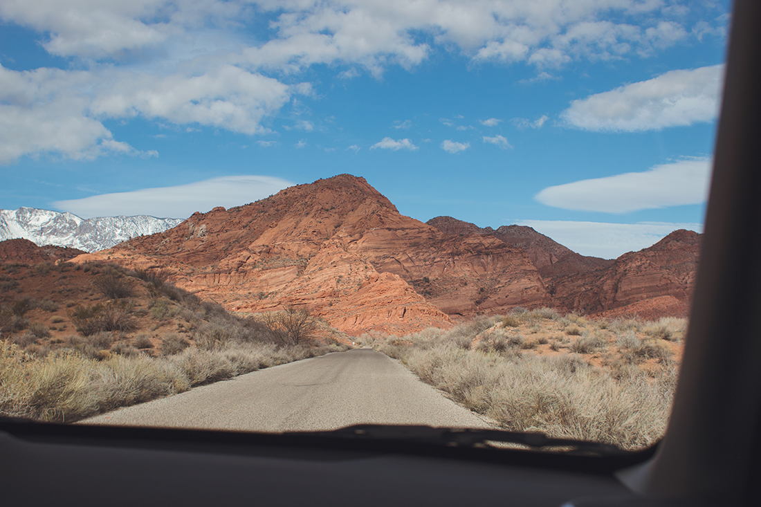 A thin, unpaved desert road leads to rugged mountains against a blue sky looming in the distance. 