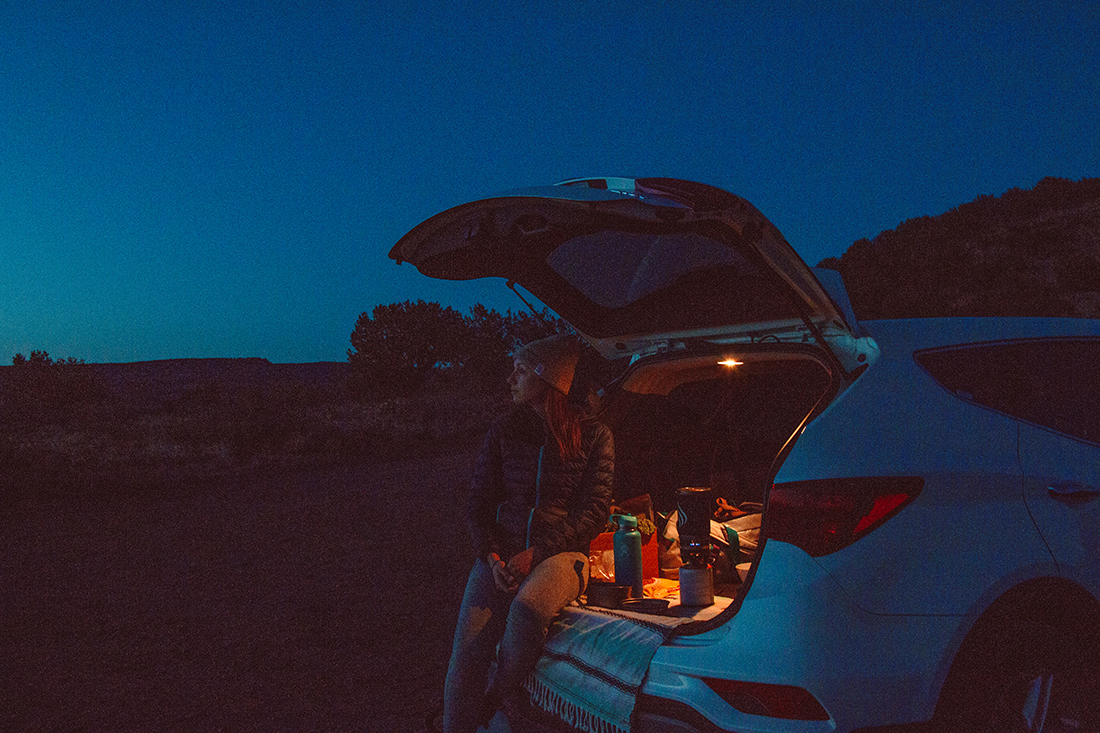 A woman gazes into the desert night as she sits in the rear of an open hatchback.