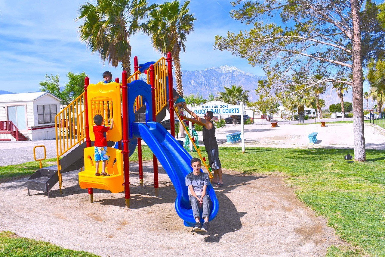 Children scamper on a colorful play structure with manufactured housing and palm trees in the background.