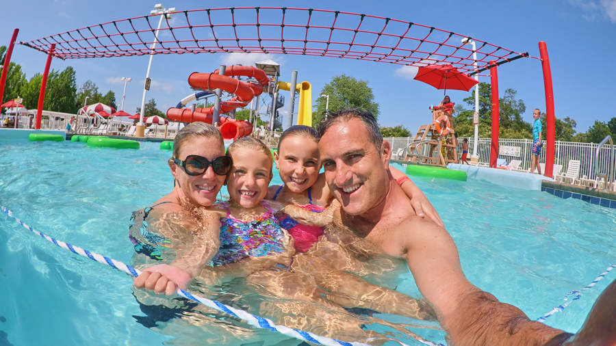 Parents and their two children pose for a selfie in a swimming pool with waterslide in the background.