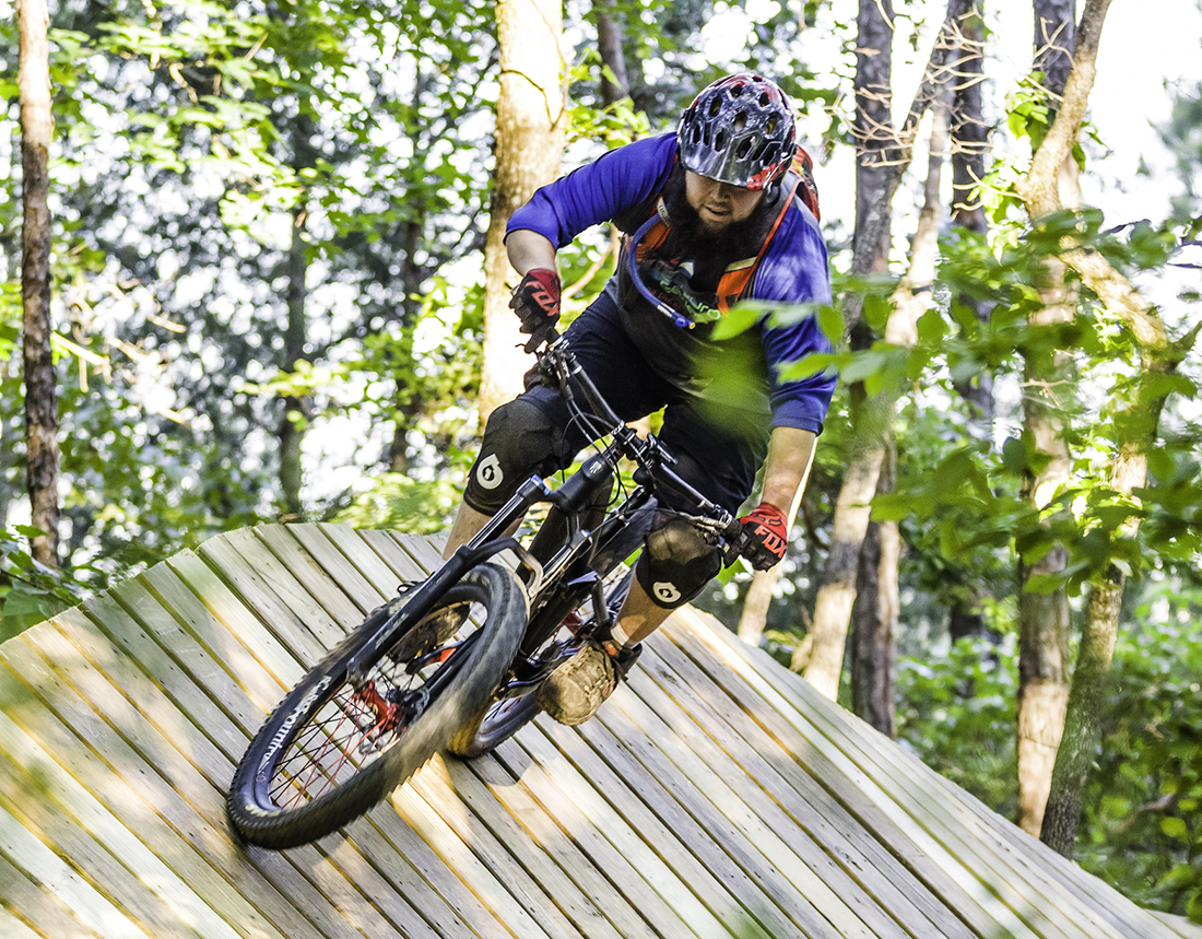 A bicycle rider takes a banked turn on a track made of wood in a forest setting.