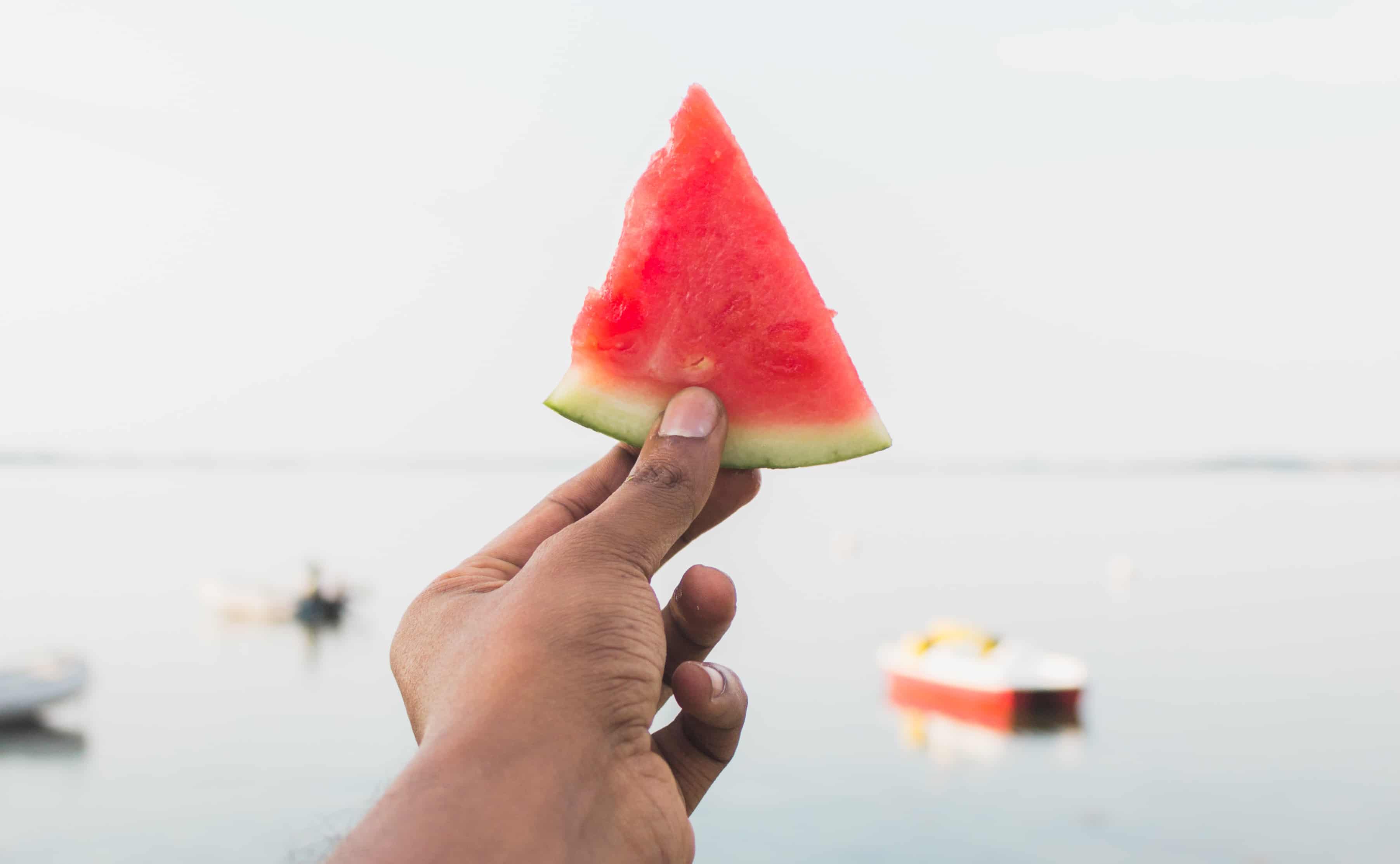 A hand holds up a triangular slice of seedless watermelon in front of a red boat. 