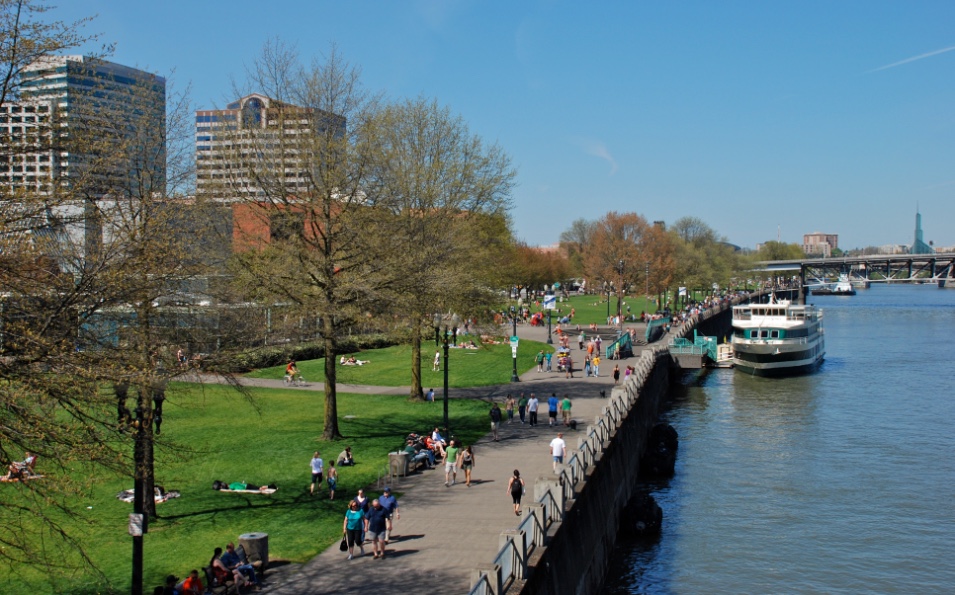 Walkers and joggers move on a riverwalk alongside a wide river with boats.