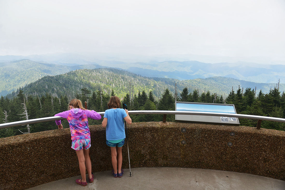 Two girls stand at a railing looking down over a mountainous landscape carpeted in forest.