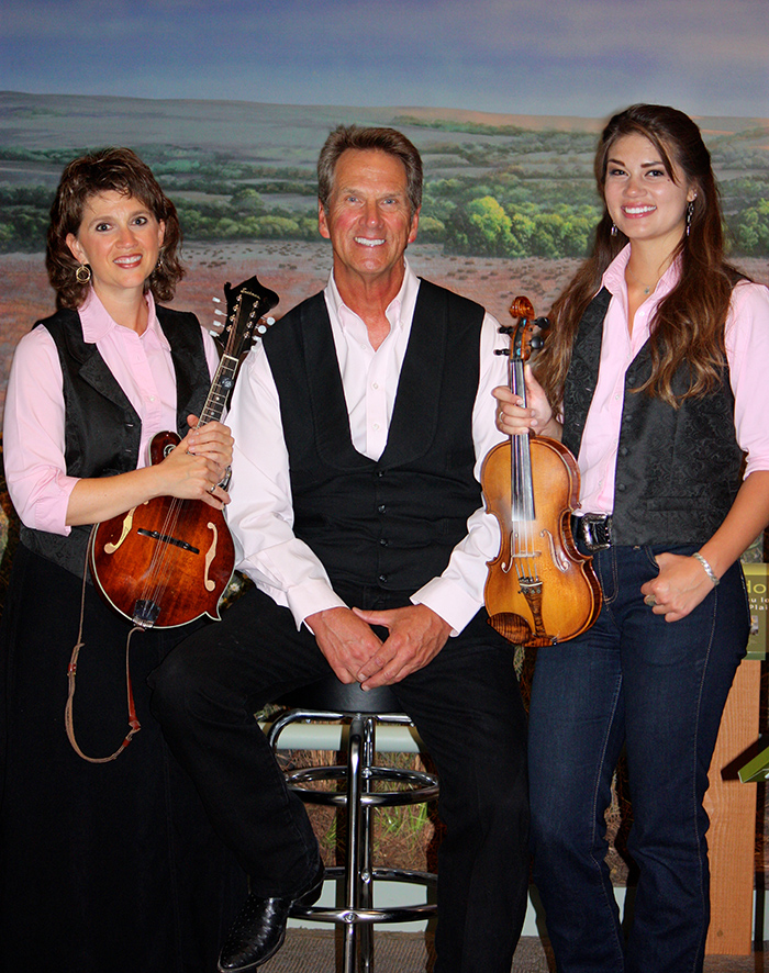 A musician in his 50s or 60s is flanked by two younger famale musicians, holding a mandolin and fiddle, respectively.