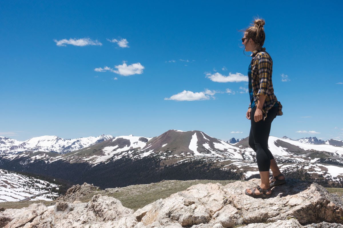 A woman walks along a rock outcropping with snow-capped peaks in the background.