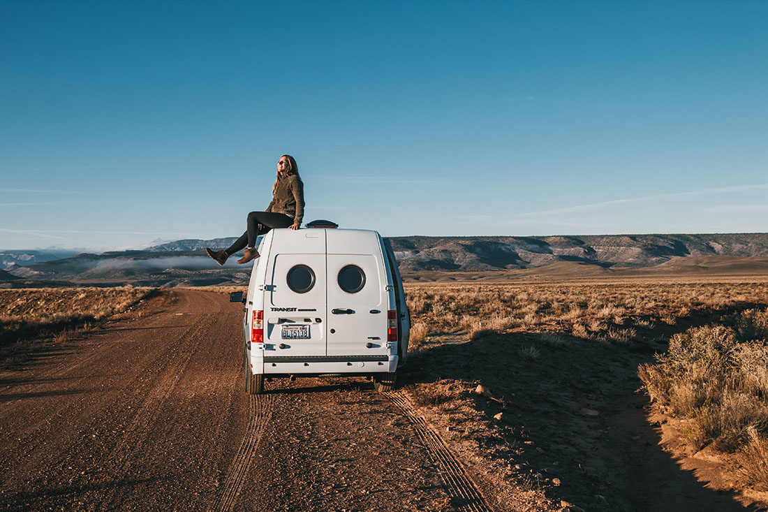 A woman wearing jacket and black pants on the roof of a camper van during sunset.