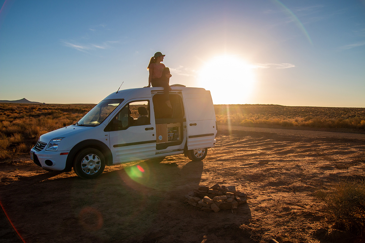 A woman sitting atop a white camper van as the sun sets on a desert horizon.