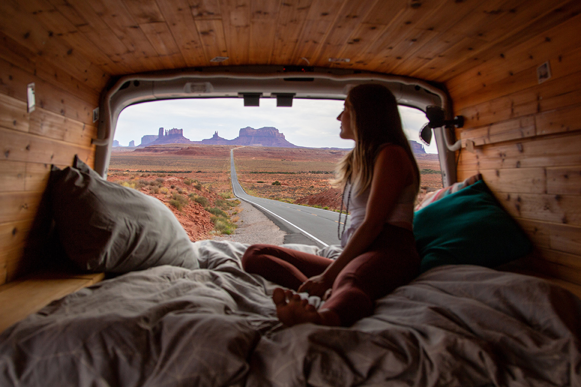 A young woman sitting in the rear of her camper van looks out at the stark mesas that line the desert horizon.