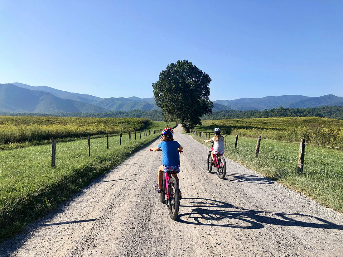 A pair of girls ride a dirt bike path toward mountains in the horizon.