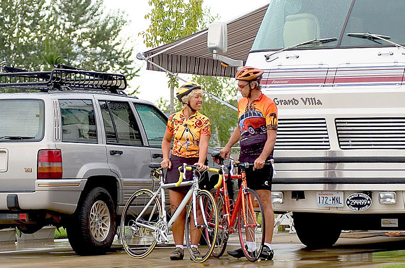 A woman and man cyclist chat as they stand next to their bikes in front of an RV.