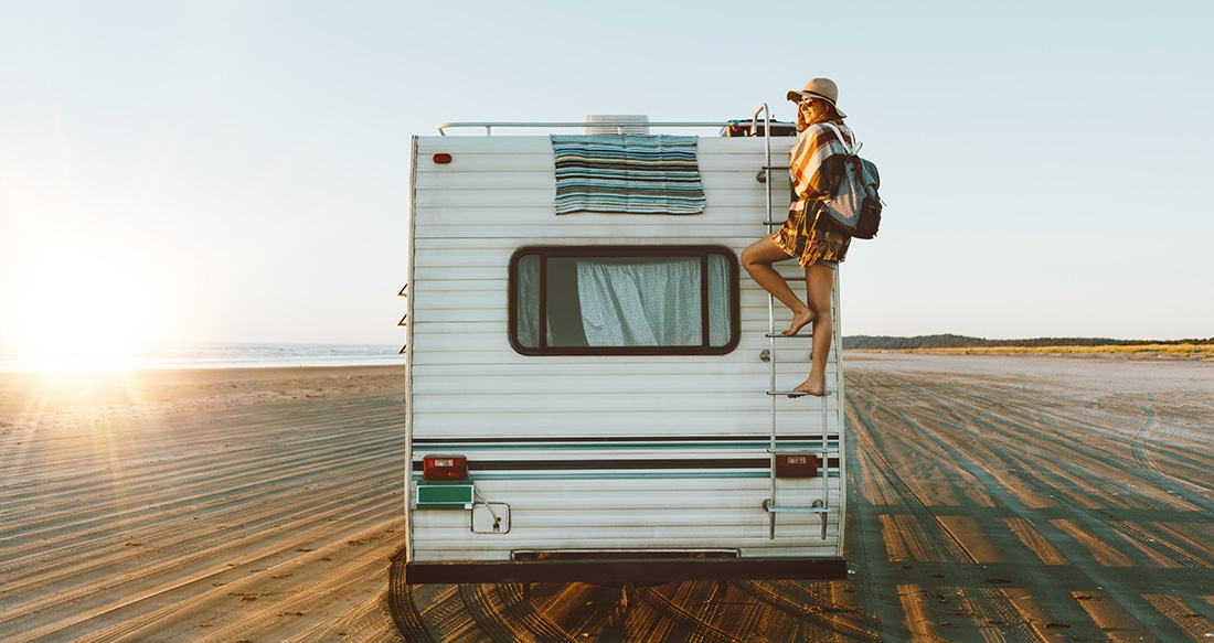 A woman in beach clothes scales the ladder of on the rear of a motorhome on an otherwise empty, sprawling beach.