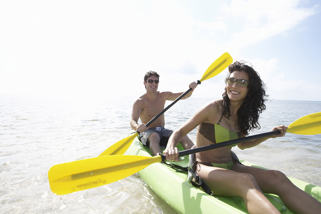 A smiling man and woman paddle a two-person green kayak with yellow paddles.