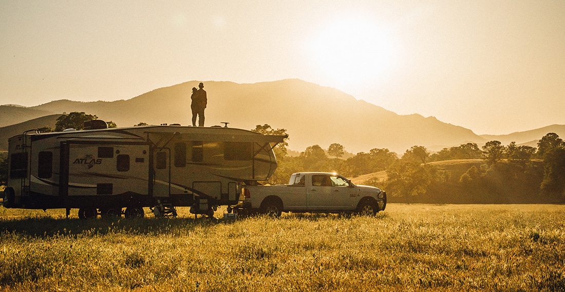 A couple stands on the roof of a fifth-wheel as the sun sets behind a mountain over a golden field.