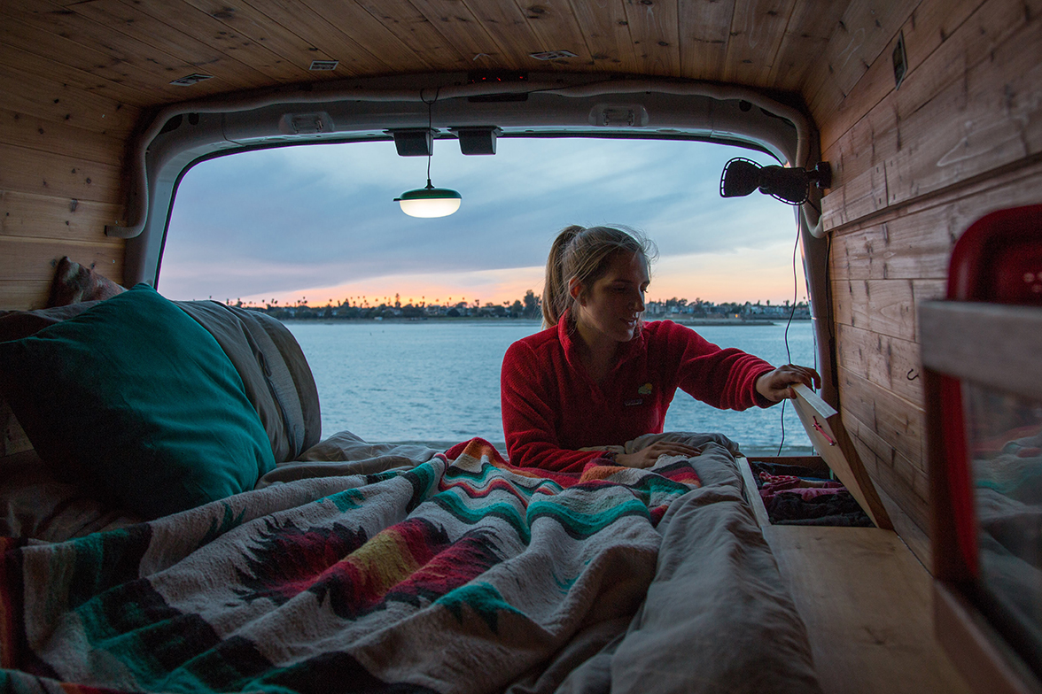 A woman inspects a storage compartment in in the back of her camper van with water in the background.