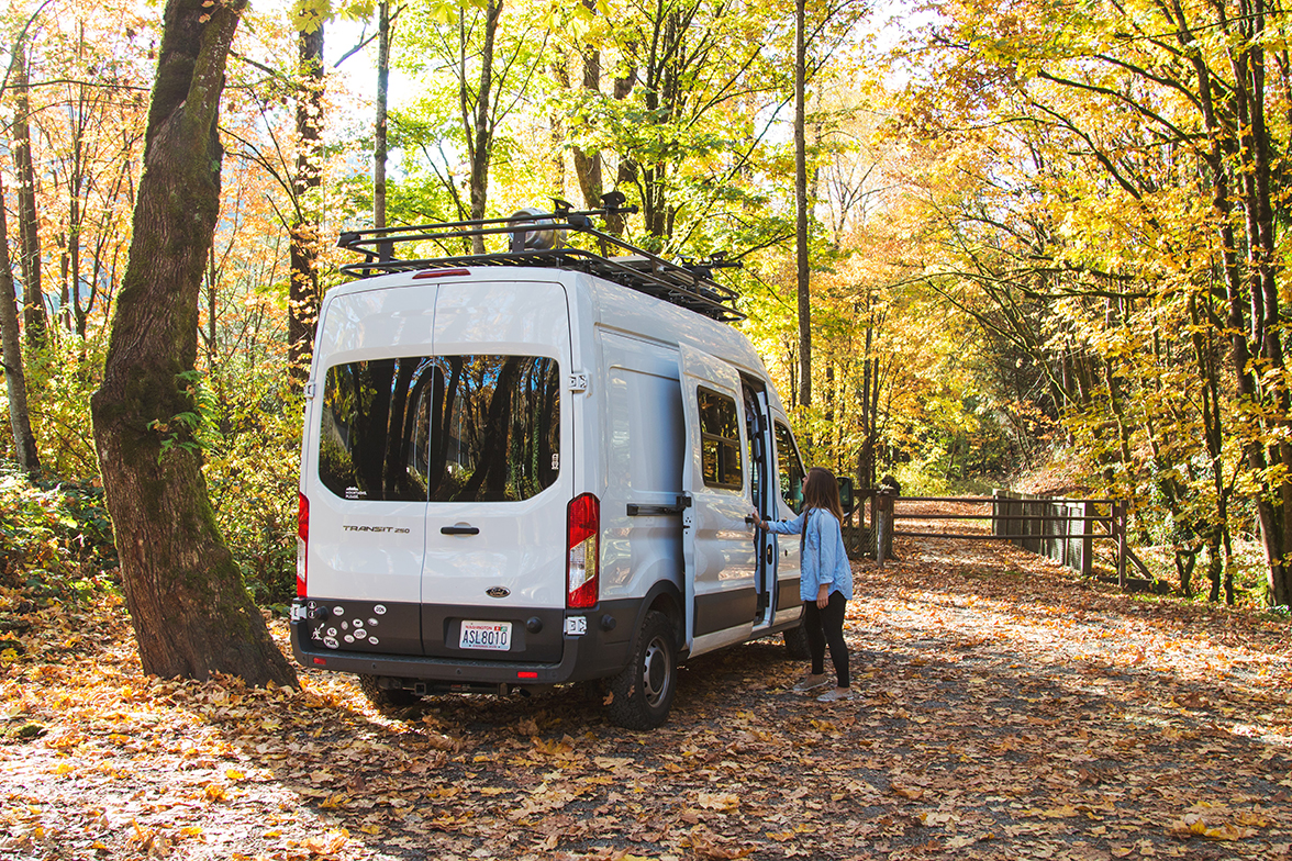 A woman opens the sliding door of her white camper van parked on leaf-covered asphalt as fall leaves adorn the trees.
