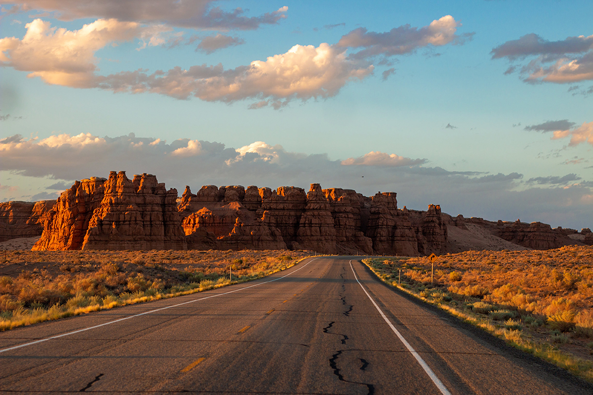 A desert highway veers right to skirt the rugged rock faces of a bluff on the horizon.