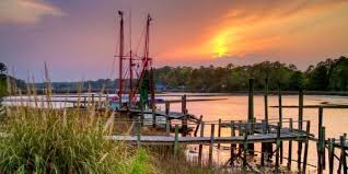 The sun sets over a wooden dock in a river or channel with a lone fishing vessel moored on serene water.