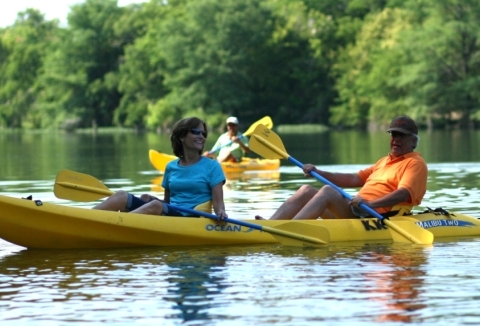 A middle-aged man and woman laugh as they paddle a yellow double kayak.