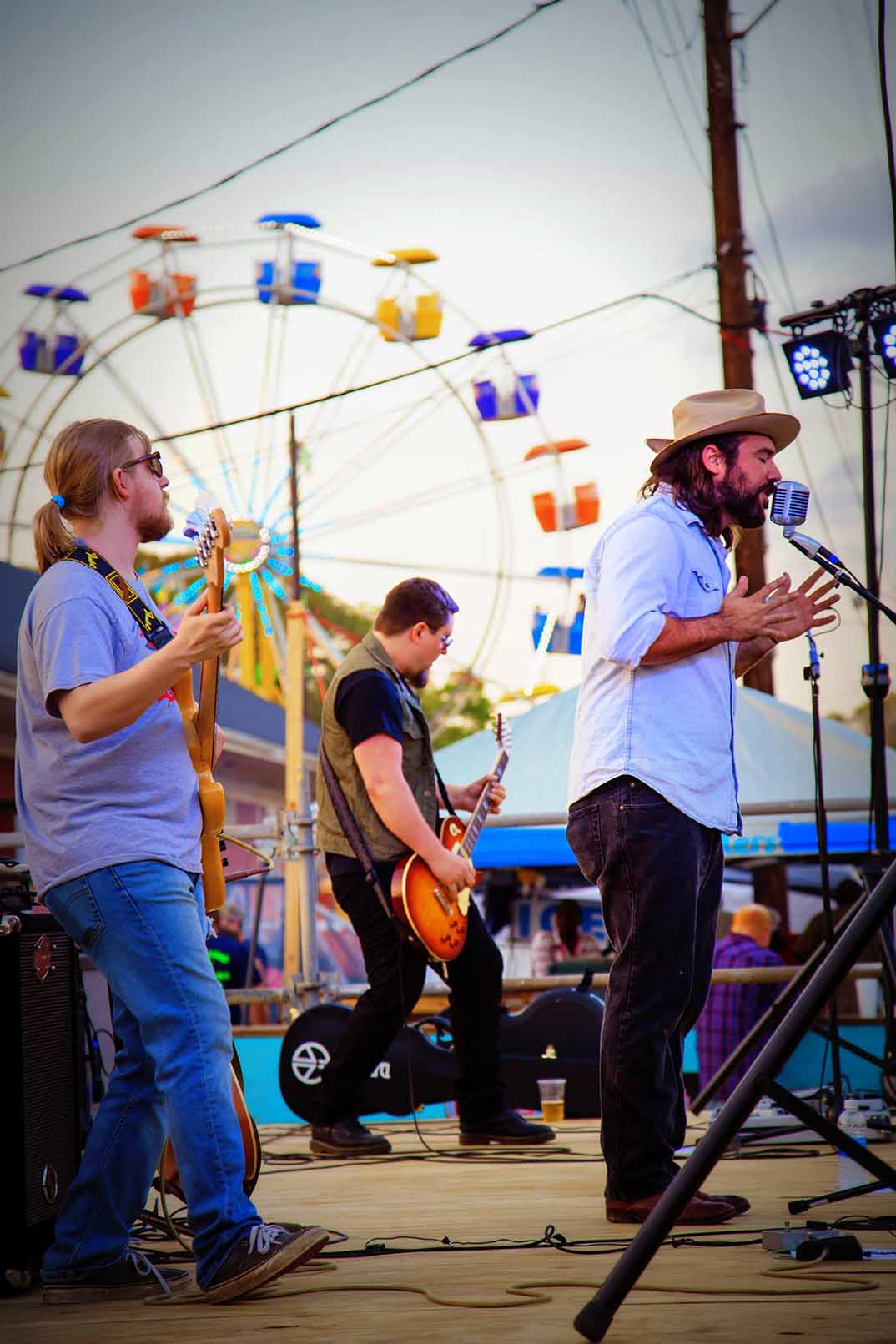 A singer in a hat with guitarist and basist and Ferris wheel in the background.