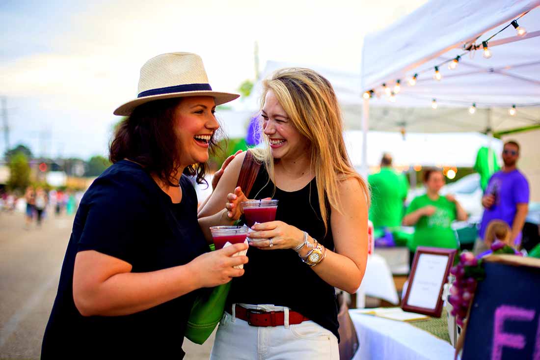 Two women share a laugh over drink as they stand near a booth at a fair or festival.