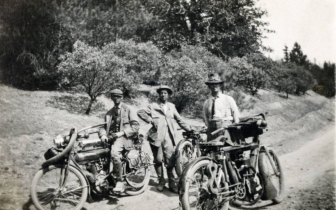 Three men standing beside old motorcycles on dirt road in the 1950s.