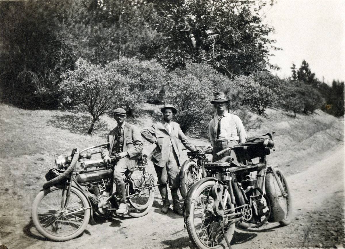 Three men standing beside old motorcycles on dirt road in the 1950s.