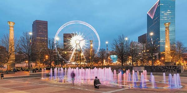 A lighted Ferris wheel spins over a park with fountains. Pillars and building fill the skyline.