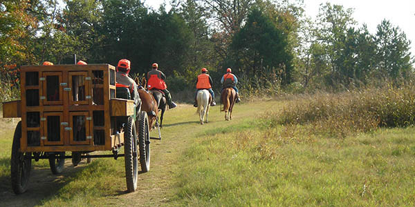 A wagon follows horseback riders down a dirt road in a hunting party.