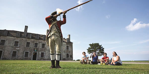 A re-enactor dressed as a British soldier fires a musket as four young people look on.