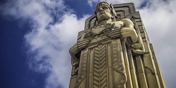 An elegant statues of a wreathed god who holds a placard with a stagecoach relief.