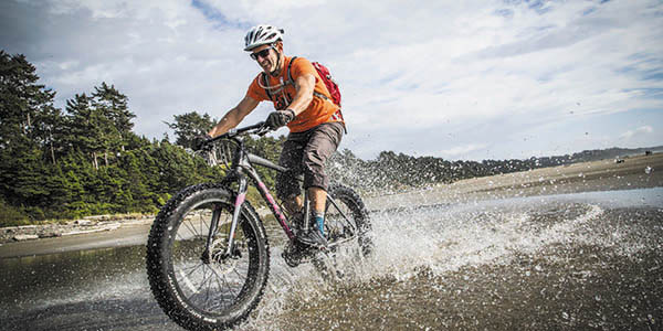 Man wearing orange shirt riding mountain bike through beach with trees in background